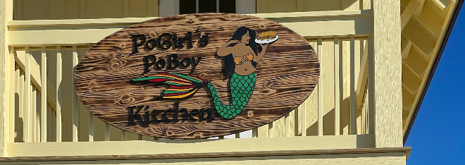 The wooden Pogirl's Poboy Kitchen sign, featuring a mermaid, on a sunny yellow porch
